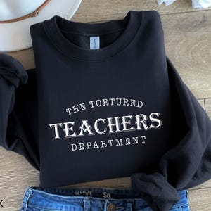 Puede incluir: Sudadera negra con el texto blanco "THE TORTURED TEACHERS DEPARTMENT". La sudadera est&aacute; doblada, con unos vaqueros azules visibles en la parte inferior. Un sombrero blanco est&aacute; en el fondo.