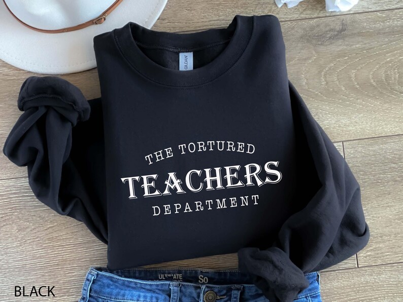 Puede incluir: Sudadera negra con el texto blanco "THE TORTURED TEACHERS DEPARTMENT". La sudadera est&aacute; doblada, con unos vaqueros azules visibles en la parte inferior. Un sombrero blanco est&aacute; en el fondo.