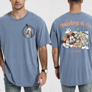 May include: A blue t-shirt featuring a cartoon of Goofy on the front and a group of Disney characters on the back. The back of the shirt has the text "Mickey & Co. - Est. 1928". The shirt is a casual style.