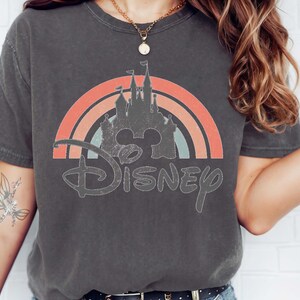 May include: Dark gray t-shirt featuring a distressed Disney castle design within a rainbow arch. The word "Disney" is written in a vintage font below the castle. The shirt is a comfortable, casual style.