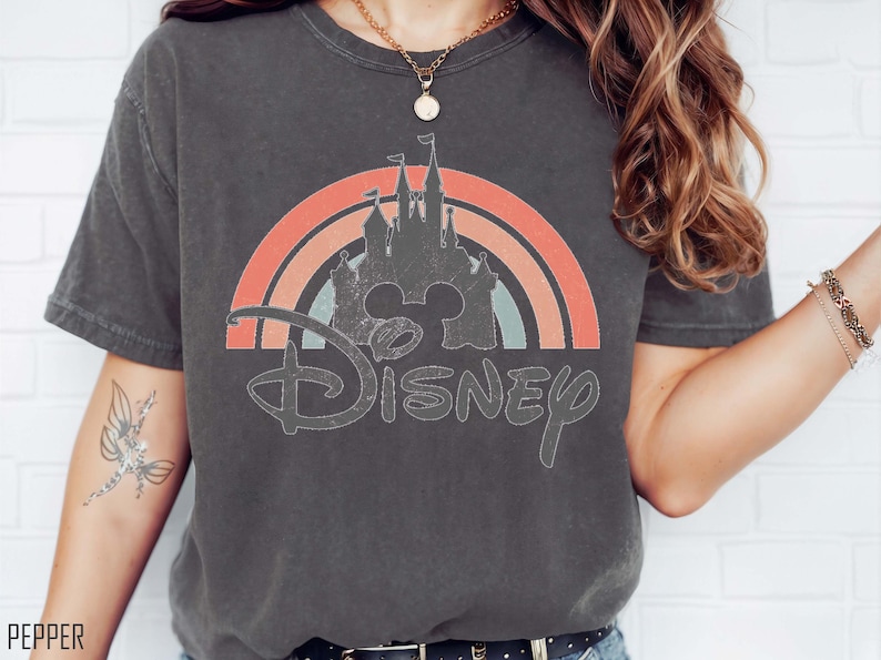 May include: Dark gray t-shirt featuring a distressed Disney castle design within a rainbow arch. The word "Disney" is written in a vintage font below the castle. The shirt is a comfortable, casual style.