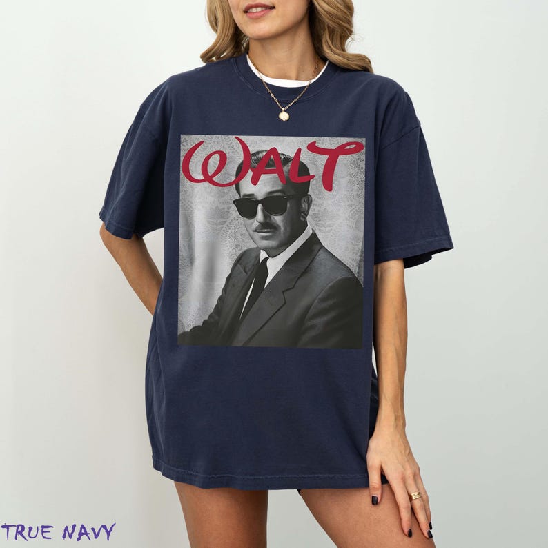 May include: A navy blue short-sleeve t-shirt featuring a black and white photographic print of a man wearing a suit and sunglasses. Above the portrait, the word WALT is printed in a bold, red, stylized script font against a light gray background.