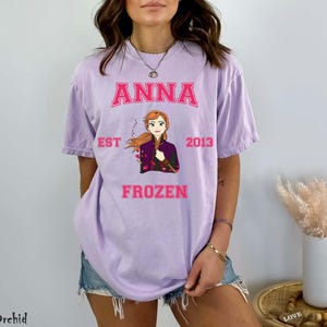 May include: A light purple t-shirt featuring the name "ANNA" in pink block letters above an illustration of a character from the movie Frozen. The text "EST 2013" and "FROZEN" are also in pink. The shirt is a casual style.