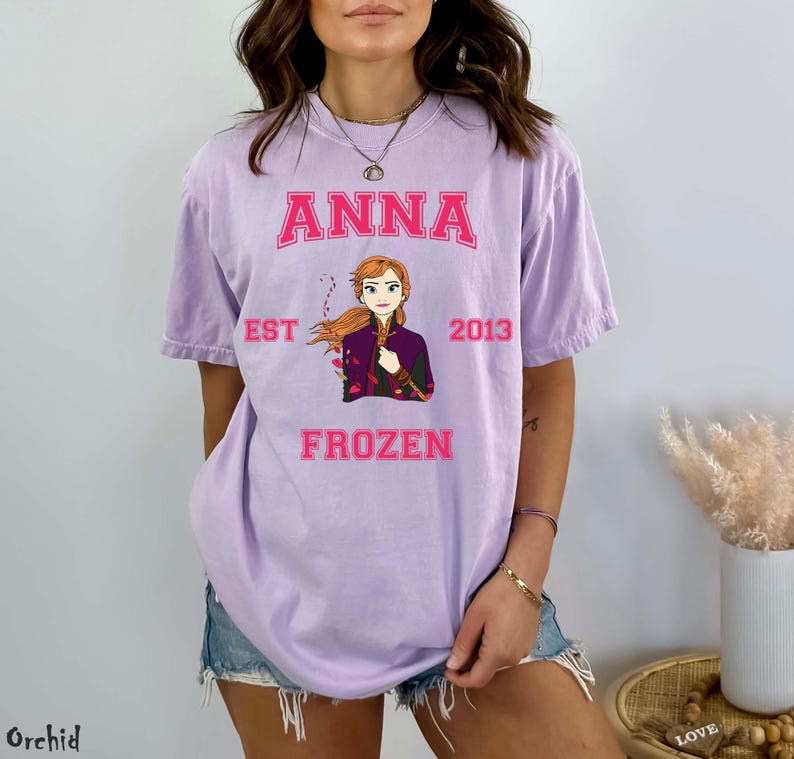 May include: A light purple t-shirt featuring the name "ANNA" in pink block letters above an illustration of a character from the movie Frozen. The text "EST 2013" and "FROZEN" are also in pink. The shirt is a casual style.