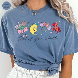 K&ouml;nnte beinhalten: Blaues Jeans-T-Shirt mit einer bunten Grafik von Zeichentrickfiguren und dem Text "Part of your World". Das Design umfasst einen Fisch, Stirnb&auml;nder und eine Krabbe. Das Shirt ist ein bequemer, l&auml;ssiger Stil.