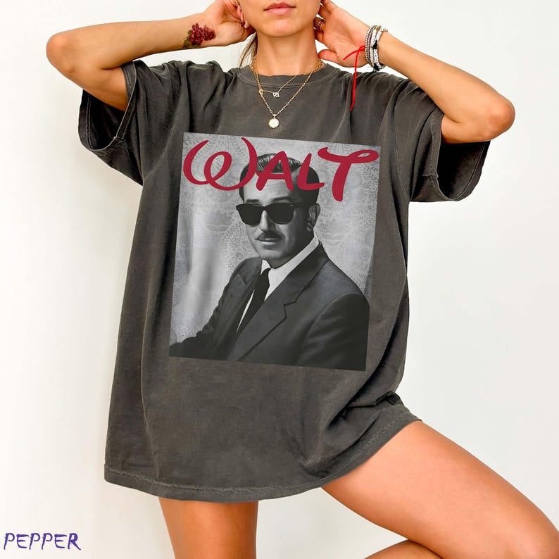 May include: A charcoal gray short-sleeve t-shirt featuring a black and white graphic print of a man in a suit and sunglasses. The word WALT is printed in large red script lettering above the portrait. The shirt has a relaxed, oversized fit.