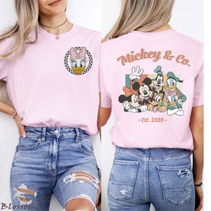 May include: A light pink t-shirt featuring Daisy Duck in a black and white checkered circle on the front. The back of the shirt displays Mickey & Co. with Mickey Mouse, Minnie Mouse, Goofy, Donald Duck, and Pluto, with "-Est. 1928-" below.