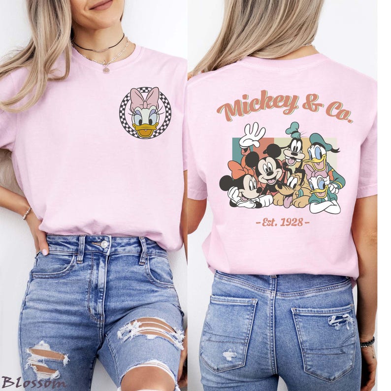 May include: A light pink t-shirt featuring Daisy Duck in a black and white checkered circle on the front. The back of the shirt displays Mickey & Co. with Mickey Mouse, Minnie Mouse, Goofy, Donald Duck, and Pluto, with "-Est. 1928-" below.