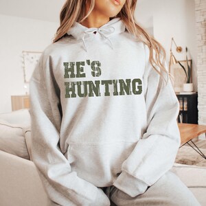 May include: A light gray hoodie with the text "HE'S HUNTING" in distressed green lettering. The hoodie has a drawstring hood and a front pocket. The sweatshirt is part of a clothing collection.