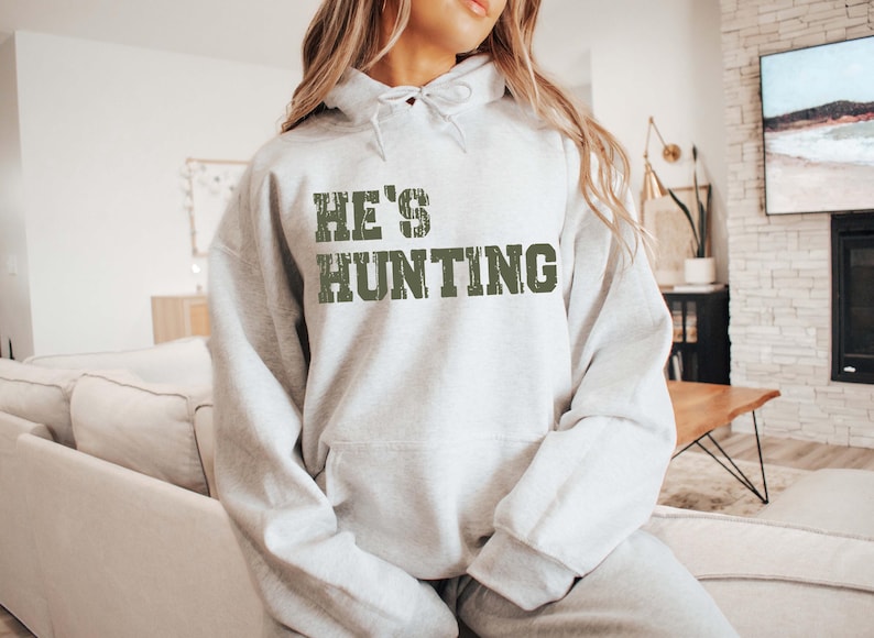 May include: A light gray hoodie with the text "HE'S HUNTING" in distressed green lettering. The hoodie has a drawstring hood and a front pocket. The sweatshirt is part of a clothing collection.
