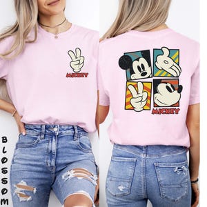 May include: Pale pink t-shirt with a Mickey Mouse graphic. The front shows a peace sign made with Mickey's hand and the word "Mickey". The back has a four-square design with Mickey's face and hand. The word "Mickey" is also printed on the back.