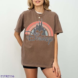May include: Brown t-shirt featuring a rainbow design with a castle silhouette and the word "Disney". The shirt has a vintage, distressed look. The word "Espresso" is printed at the bottom left.