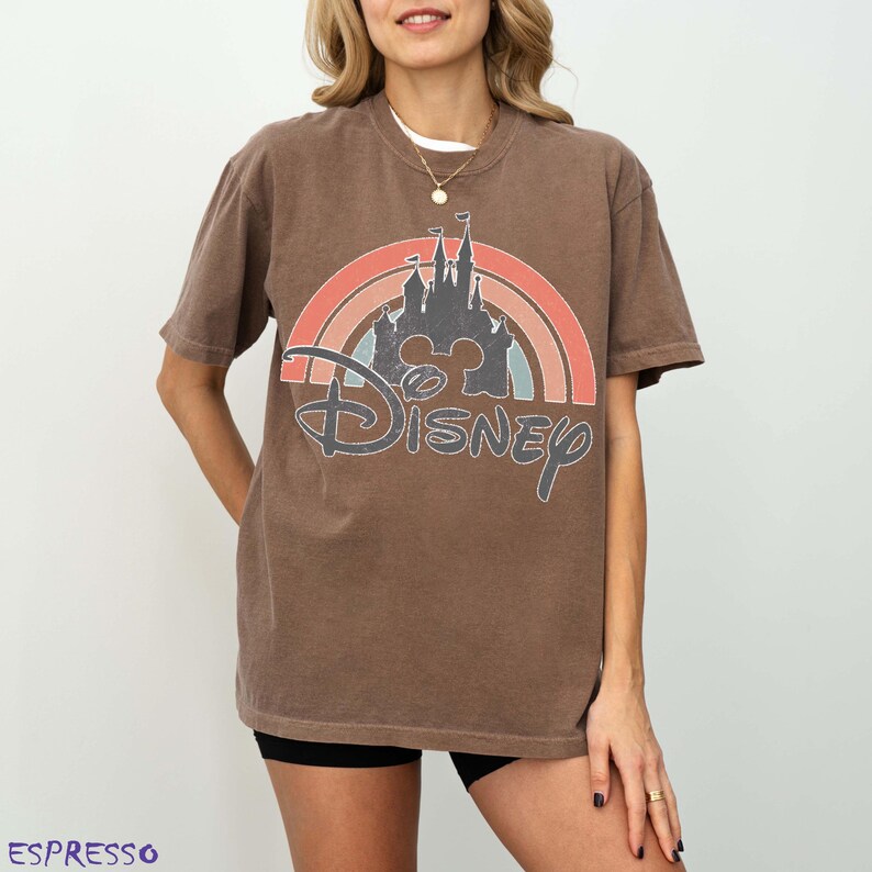 May include: Brown t-shirt featuring a rainbow design with a castle silhouette and the word "Disney". The shirt has a vintage, distressed look. The word "Espresso" is printed at the bottom left.