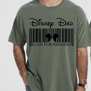 K&ouml;nnte beinhalten: Olivgr&uuml;nes T-Shirt mit dem Text "Disney Dad" &uuml;ber einem Barcode-Design. Der Barcode enth&auml;lt eine Silhouette von Mickey Mouse-Ohren und den Text "SCAN FOR PAYMENT".