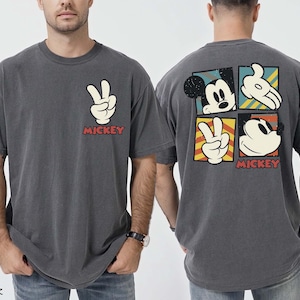 May include: Dark grey t-shirt with a white peace sign and the word "MICKEY" in red. The back features Mickey Mouse graphics in a square grid. A casual, vintage-style tee, suitable for everyday wear.