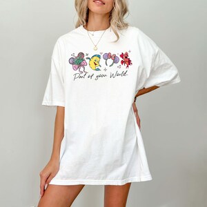 K&ouml;nnte beinhalten: Wei&szlig;es T-Shirt mit einem farbenfrohen Grafikdesign und dem Text "Part of your World". Die Grafik zeigt Cartoon-Bilder einer Meereskreatur, eines gelben Fisches und einer roten Meereskreatur. Das T-Shirt hat kurze &Auml;rmel und eine lockere Passform.