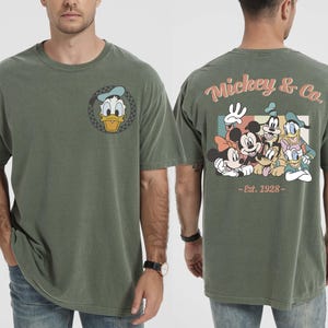 May include: Olive green t-shirt featuring Donald Duck on the front and a group of Disney characters on the back. The back of the shirt has the text "Mickey & Co. - Est. 1928". The shirt is a short-sleeved, crew neck style.