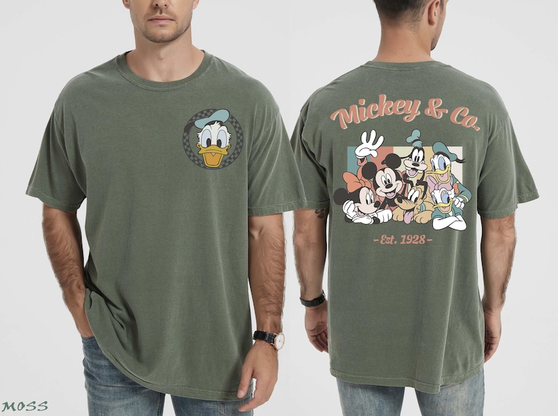 May include: Olive green t-shirt featuring Donald Duck on the front and a group of Disney characters on the back. The back of the shirt has the text "Mickey & Co. - Est. 1928". The shirt is a short-sleeved, crew neck style.