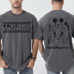 May include: A charcoal gray short-sleeve t-shirt featuring a black checkered band with Disney character faces on the front and a larger grid pattern with character faces and the text Walt Disney World on the back. The shirt is labeled as pepper color.