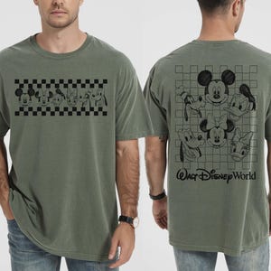 May include: An olive green short-sleeve t-shirt featuring a black graphic of Disney character faces on a checkered band on the front, and a larger grid graphic with character faces and the text Walt Disney World on the back.