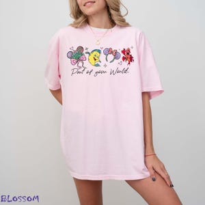 K&ouml;nnte beinhalten: Hellrosa T-Shirt mit einem Aufdruck, der eine Schleife, eine gelbe Figur und Ohren zeigt. Der Text "Part of your World" ist darunter gedruckt. Das T-Shirt hat kurze &Auml;rmel und eine &uuml;bergro&szlig;e Passform.