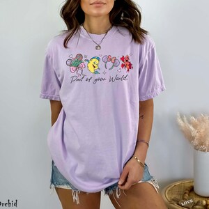 K&ouml;nnte beinhalten: Ein helllila T-Shirt mit einer Cartoon-Grafik und dem Text "Part of your World". Das Design umfasst Schleifen, Herzen und Blumen. Das Shirt hat eine lockere Passform.