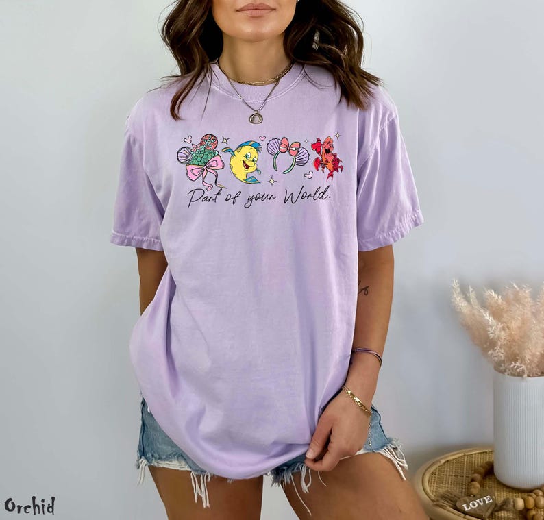 K&ouml;nnte beinhalten: Ein helllila T-Shirt mit einer Cartoon-Grafik und dem Text "Part of your World". Das Design umfasst Schleifen, Herzen und Blumen. Das Shirt hat eine lockere Passform.