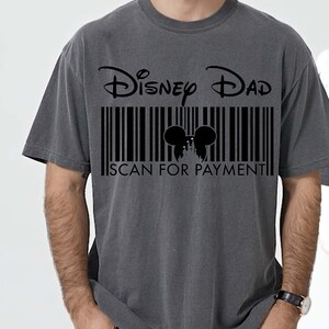 K&ouml;nnte beinhalten: Dunkelgraues T-Shirt mit der Aufschrift "Disney Dad" &uuml;ber einem schwarzen Barcode-Design. Der Barcode enth&auml;lt eine Silhouette von Mickey Mouse-Ohren und den Text "SCAN FOR PAYMENT".