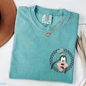 May include: Seafoam green t-shirt featuring a cartoon graphic of Goofy in a chef's hat within a checkered circle. The shirt has a crew neck and short sleeves. A gold necklace with a pendant is draped across the shirt.