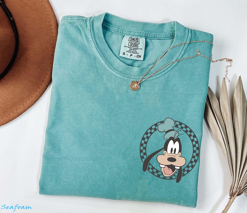 May include: Seafoam green t-shirt featuring a cartoon graphic of Goofy in a chef's hat within a checkered circle. The shirt has a crew neck and short sleeves. A gold necklace with a pendant is draped across the shirt.