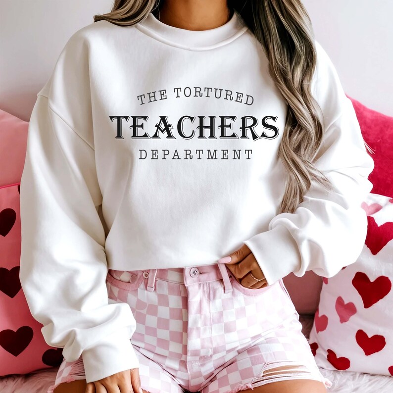 Puede incluir: Una sudadera blanca con el texto "THE TORTURED TEACHERS DEPARTMENT" en negro. La persona lleva unos pantalones cortos a cuadros rosas y blancos. El fondo incluye cojines rosas con dise&ntilde;os de coraz&oacute;n.