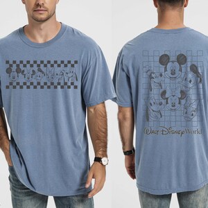 May include: A blue jean colored t-shirt featuring a front graphic of Disney characters in a black and white checkered band. The back displays a larger grid pattern with character faces and the text Walt Disney World in a dark script font.