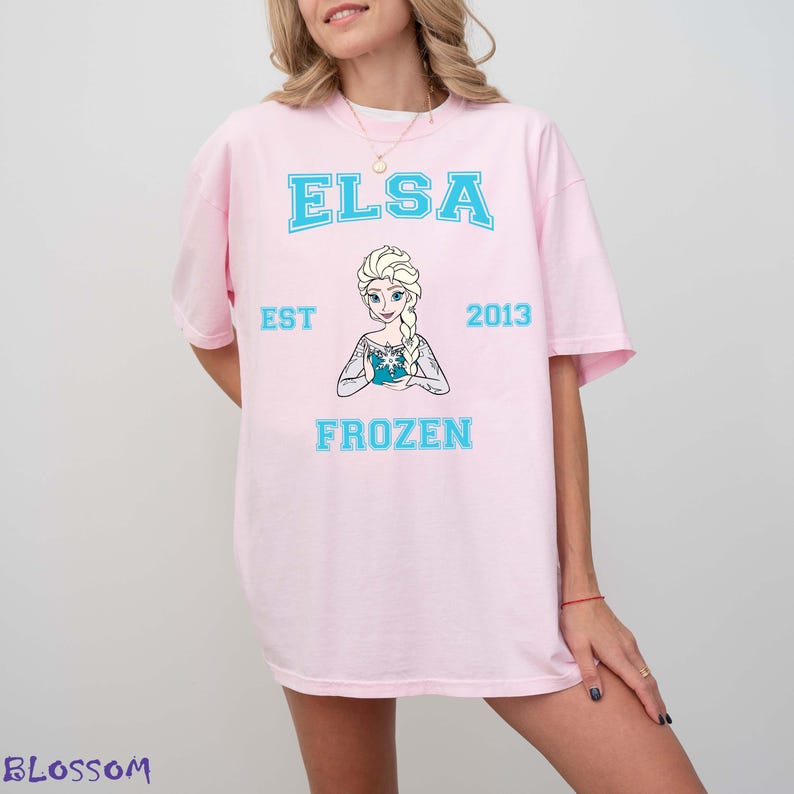 May include: A light pink t-shirt featuring the name "ELSA" in light blue block letters above a graphic of Elsa from the movie Frozen. Below the graphic are the words "EST 2013" and "FROZEN" in light blue.