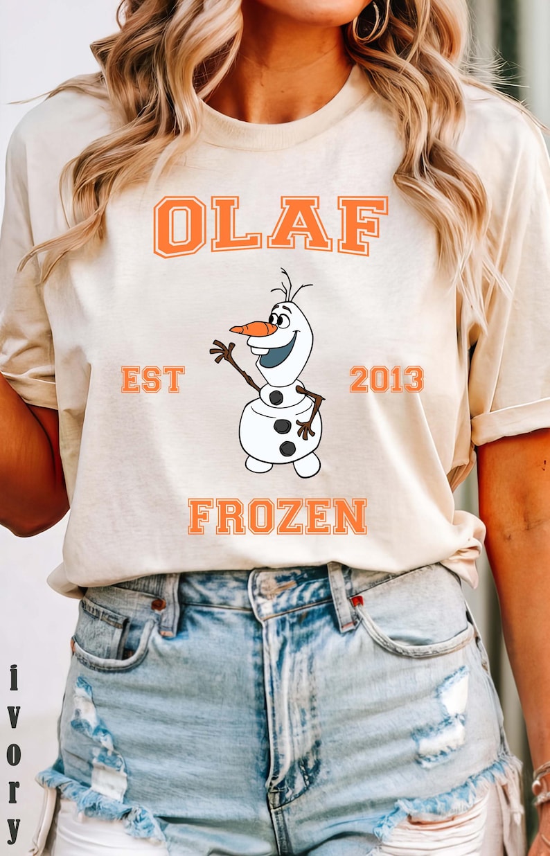 May include: A beige t-shirt featuring the word "OLAF" in orange block letters above a cartoon image of Olaf the snowman. Below the image are the words "FROZEN" and "EST 2013" in orange. The shirt is paired with denim shorts.