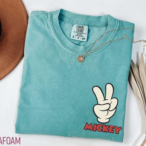 May include: Seafoam green t-shirt with a Mickey Mouse peace sign graphic. The graphic features a white hand with black outlines making a peace sign, and the word "Mickey" in red block letters. A gold necklace with a pendant is on the shirt.