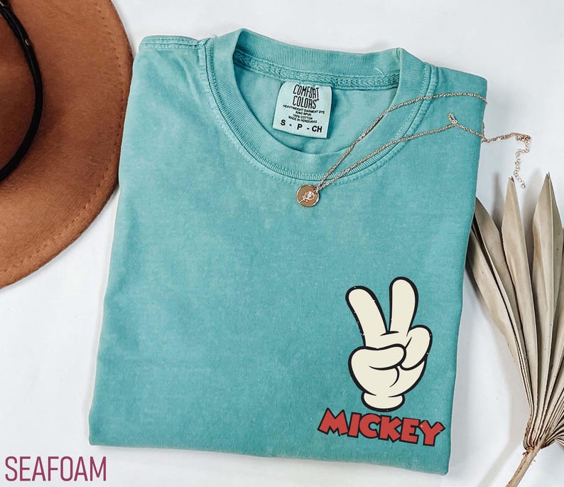 May include: Seafoam green t-shirt with a Mickey Mouse peace sign graphic. The graphic features a white hand with black outlines making a peace sign, and the word "Mickey" in red block letters. A gold necklace with a pendant is on the shirt.