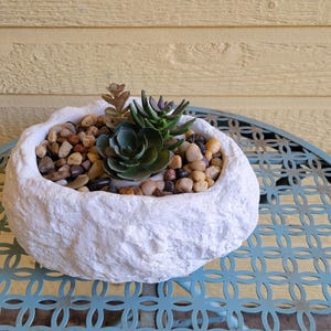 May include: A white, textured, oval-shaped planter with a succulent plant and small stones. The planter is sitting on a blue metal table.