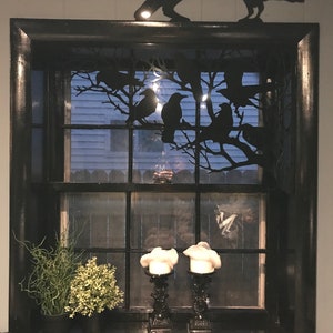May include: A black window frame with a black metal silhouette of a tree with crows perched on the branches. Two white candles are on the window sill in black candle holders.