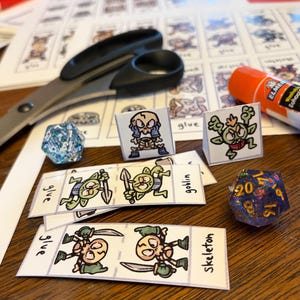 May include: A tabletop scene with crafting supplies for a tabletop game. Includes scissors, a glue stick, dice, and printed paper cutouts of goblins and skeletons. The dice are blue and purple.