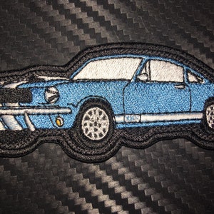 May include: A blue embroidered patch of a classic Ford Mustang car. The patch has a white stripe along the side and a black background.