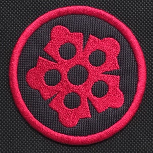 May include: A circular embroidered patch featuring a red floral design with six petals and six inner circles, set against a black textured background. The design is framed by a red border.