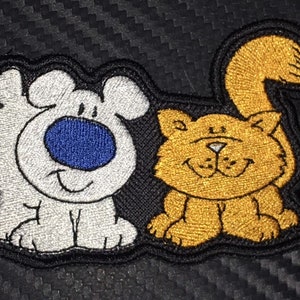 May include: Embroidered patch featuring a white dog and an orange cat. The dog is smiling and has a blue nose. The cat is also smiling and has a white nose. Both animals are sitting on a black background.