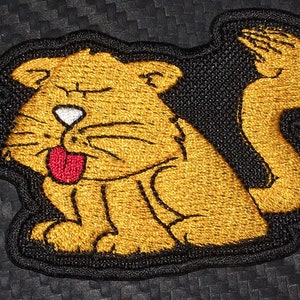 May include: Embroidered patch of a cartoon cat with a brown body, black outline, and a red tongue sticking out. The cat is sitting with its tail curled up behind it.