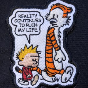 May include: Embroidered patch featuring Calvin and Hobbes. Calvin, with spiky yellow hair and a red jacket, walks alongside Hobbes, an orange and black tiger. A speech bubble reads, "REALITY CONTINUES TO RUIN MY LIFE."