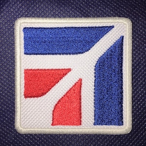 May include: White embroidered patch with a blue and red geometric design. The design features a blue rectangle with a red triangle and a red trapezoid.