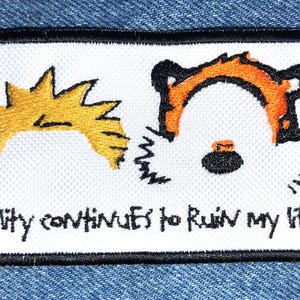 May include: White embroidered patch with a cartoon image of Calvin and Hobbes. Calvin is blonde with spiky hair and Hobbes is an orange tiger. The text "Reality continues to ruin my life." is embroidered below the image.