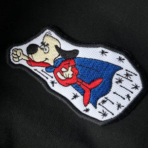 May include: A white, red, and blue embroidered patch of a cartoon dog wearing a cape and flying through the air. The dog is wearing a red cape with a blue lining and has a white background with black stars and lines.