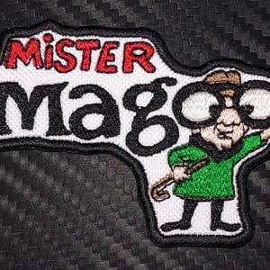 May include: White embroidered patch with the text "Mister Mago" in black and red lettering. A cartoon character with glasses, a green shirt, and a cane is depicted on the right side of the patch.