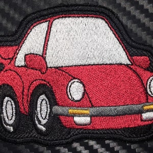 May include: Red embroidered patch of a cartoon car with black wheels and white headlights. The car has a white windshield and a black outline.