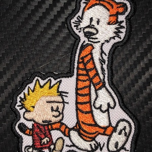 May include: An embroidered patch featuring the cartoon characters Calvin and Hobbes. Calvin is wearing a red shirt and is walking next to Hobbes, who is a tiger wearing an orange and black striped shirt.